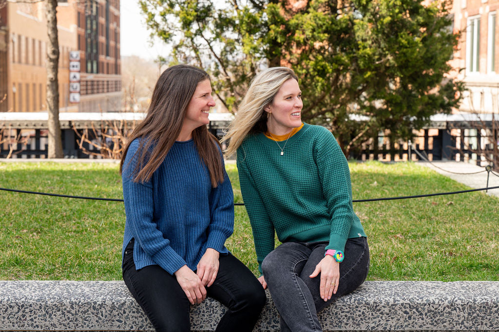 Two women sitting on a ledge outdoors, smiling and talking, wearing USA made wool sweaters by Blythly