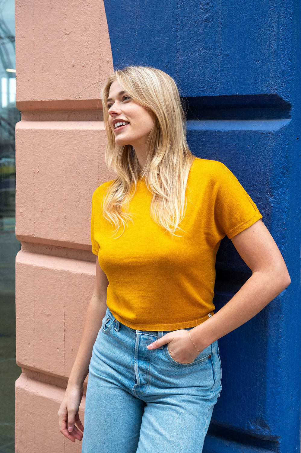 Woman in a yellow Blythly sweater tee and blue jeans standing against a blue wall.