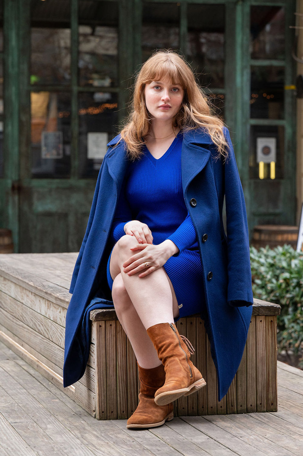 Woman in a blue coat and blue wool USA made dress by Blythly sitting on a wooden bench outdoors.