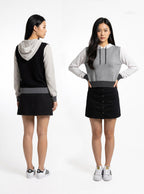 A model is shown from both the front and back wearing a color-blocked hoodie vest. The vest features a black body, a light gray sleeve and hood, and a dark gray waistband, paired with a black mini skirt and white sneakers.