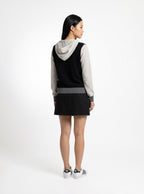 A woman is shown from the back wearing a color-blocked hooded sweatshirt dress. The dress features a black body, a light gray sleeve and hood, and a darker gray horizontal stripe at the waist.