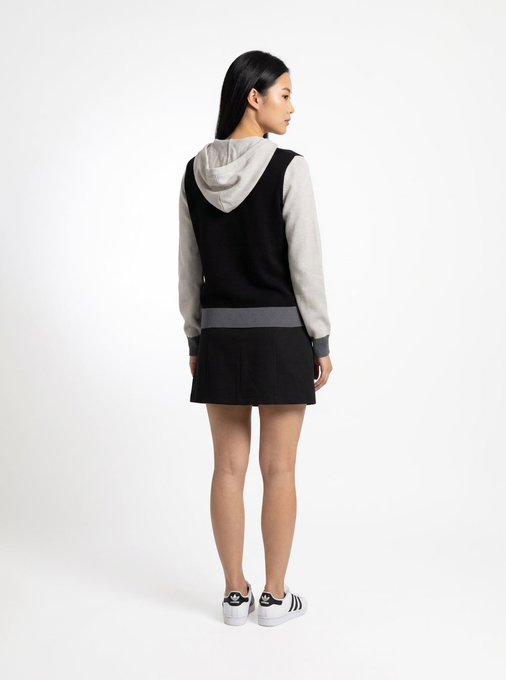 A woman is shown from the back wearing a color-blocked hooded sweatshirt dress. The dress features a black body, a light gray sleeve and hood, and a darker gray horizontal stripe at the waist.