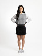 A woman modeling a sleeveless gray and white color-block hoodie with a black button-up mini skirt and white sneakers, standing against a plain white studio background.
