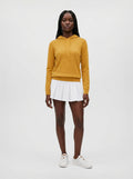 A model stands against a light gray studio background wearing a mustard yellow long-sleeve hoodie, a white pleated mini skirt, and white sneakers.